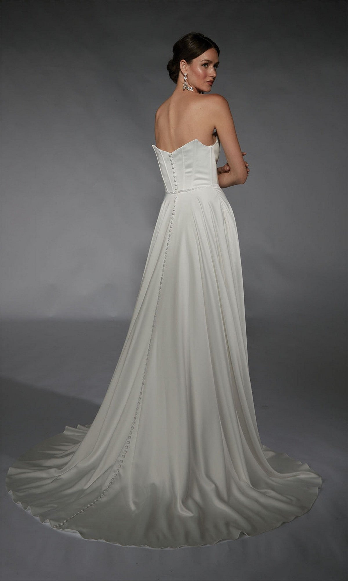 A woman poses elegantly in a studio with a gray background, wearing the Alyce 7110: a long white strapless formal gown with button details and a train.